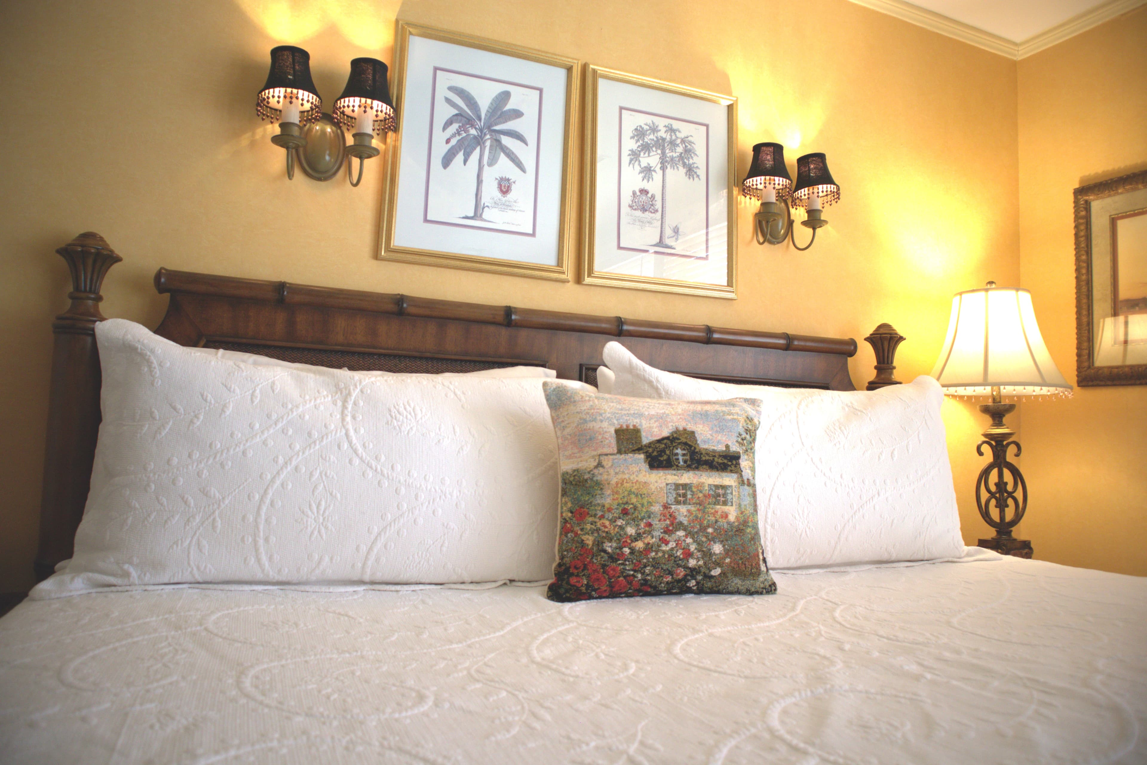 A cozy bed with white bedding and decorative pillows, flanked by wall sconces and framed artwork.