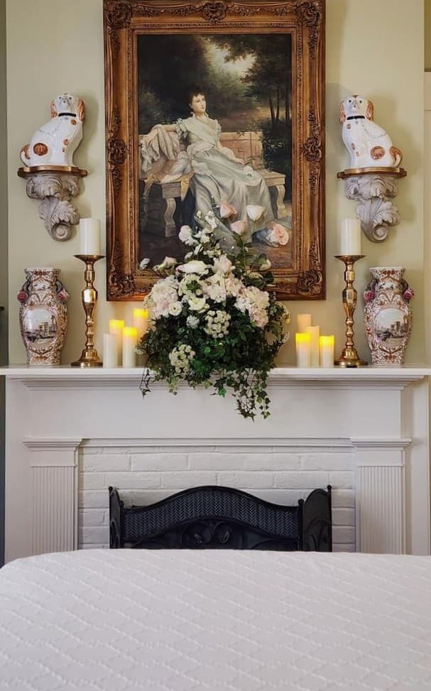 A vintage-style fireplace adorned with a large portrait, floral arrangement, candles, and decorative vases.
