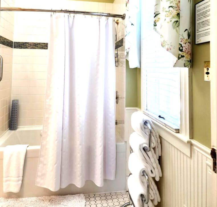 A bright, clean bathroom featuring a white shower curtain, neatly stacked towels, and floral window treatments.
