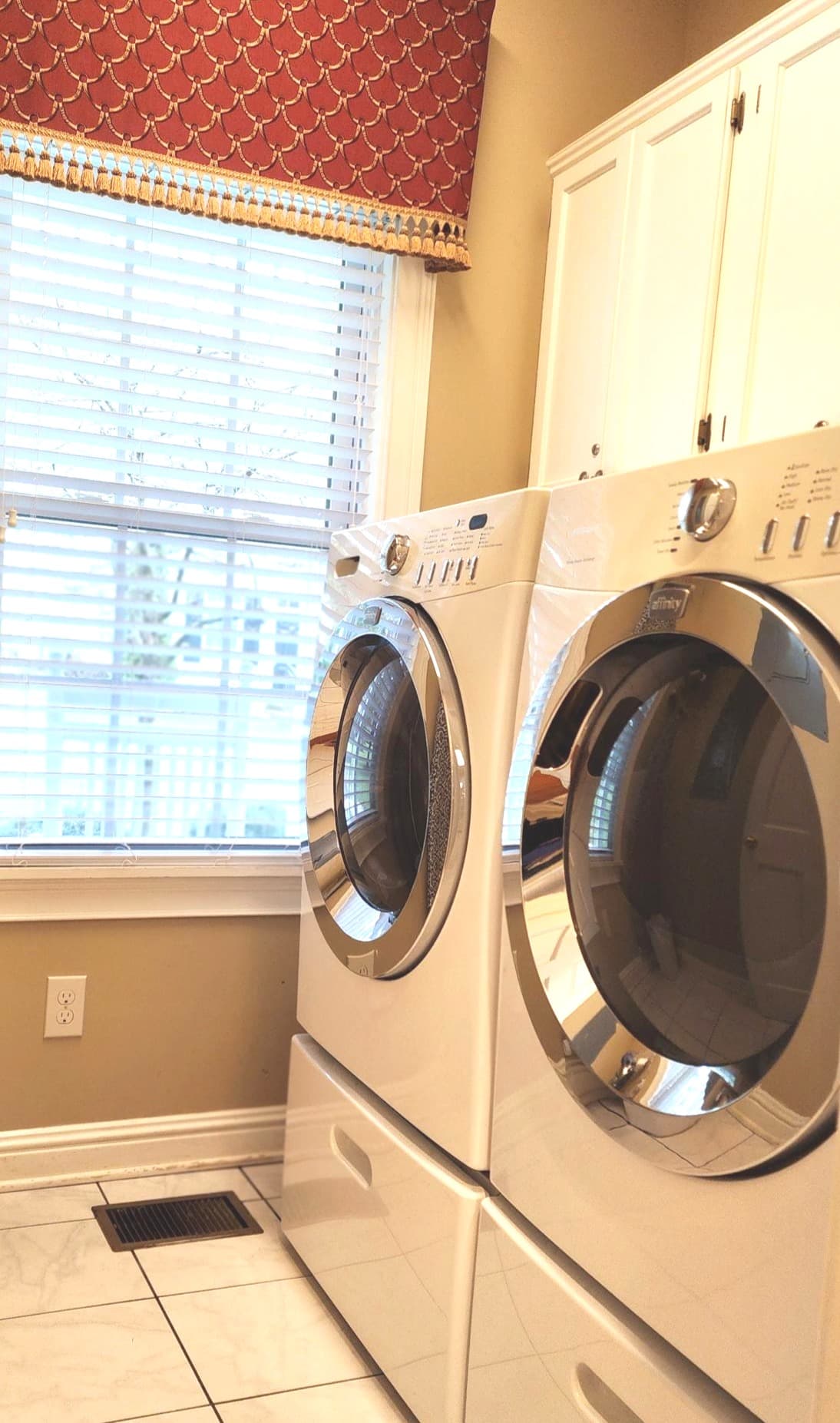 A laundry room featuring white washing machine and dryer stacked beside a window with blinds and a red patterned valance.
