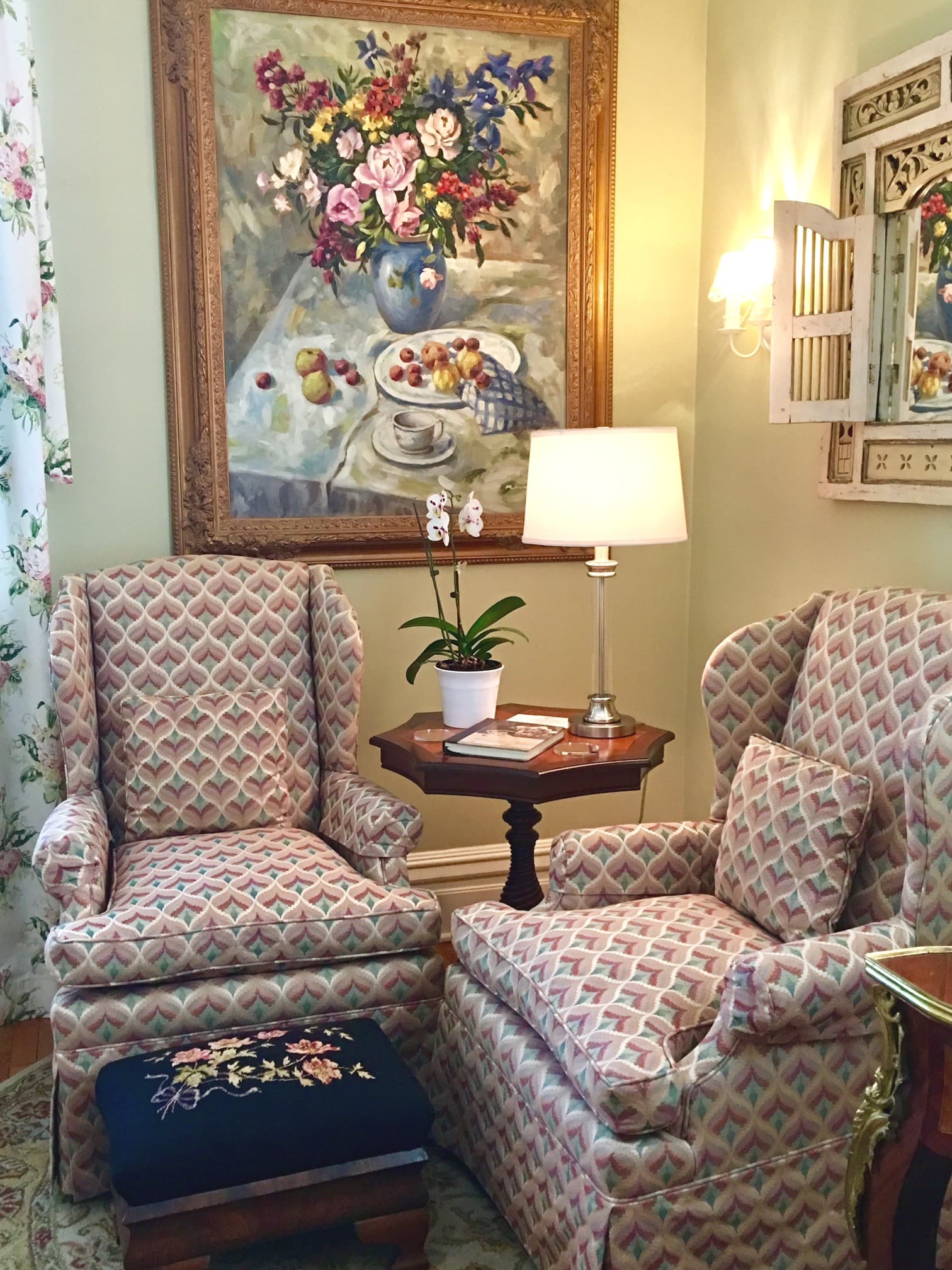 A cozy living room corner featuring patterned armchairs, a wooden side table with a lamp, and a floral painting above.