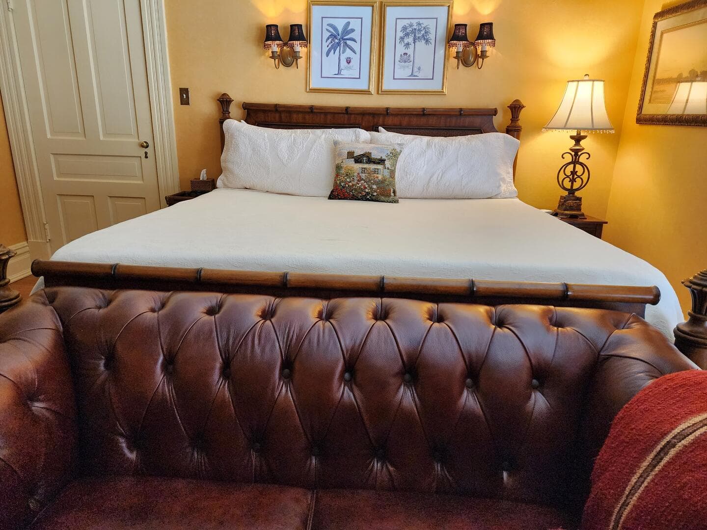 A cozy bedroom featuring a large bed with white linens, framed botanical prints on the wall, and a brown tufted leather couch in the foreground.