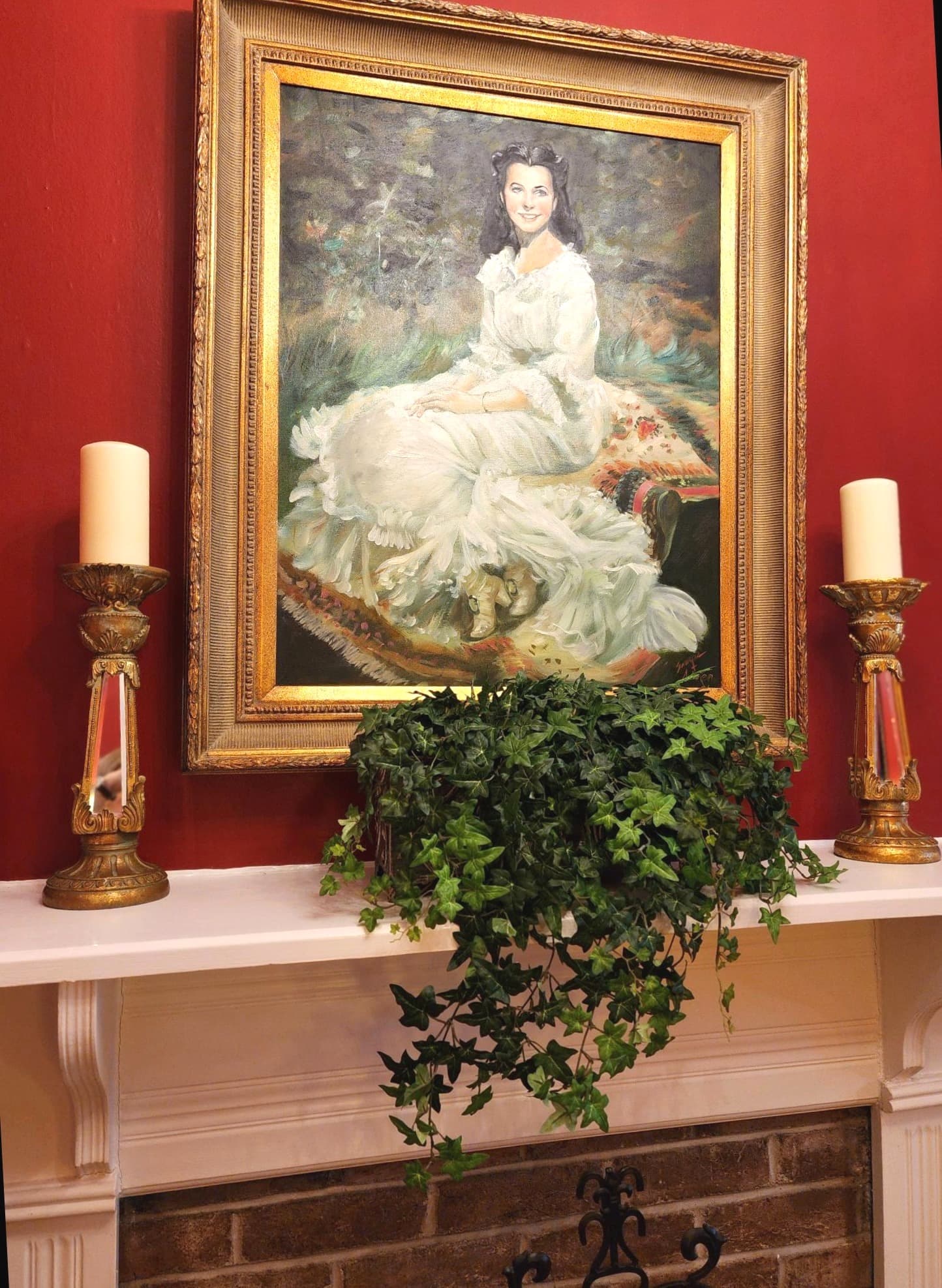 A vintage portrait of a woman in a white dress, framed and hanging above a decorative fireplace with candles and greenery.
