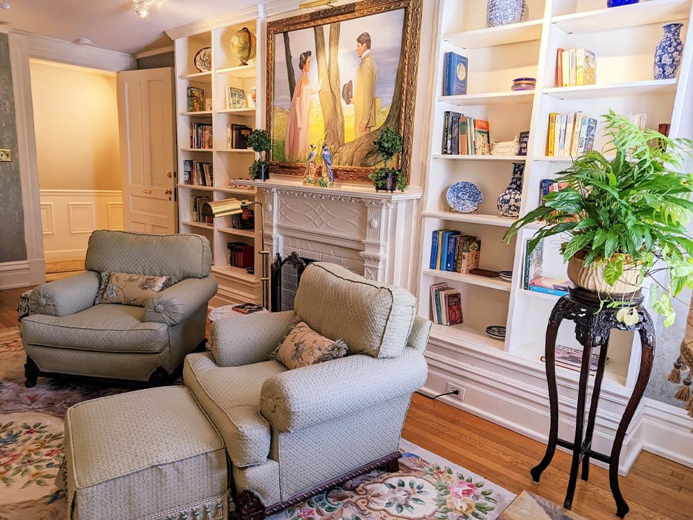 A cozy living room featuring two upholstered chairs, a decorative plant, and a bookshelf filled with books, all under a painting of two figures.