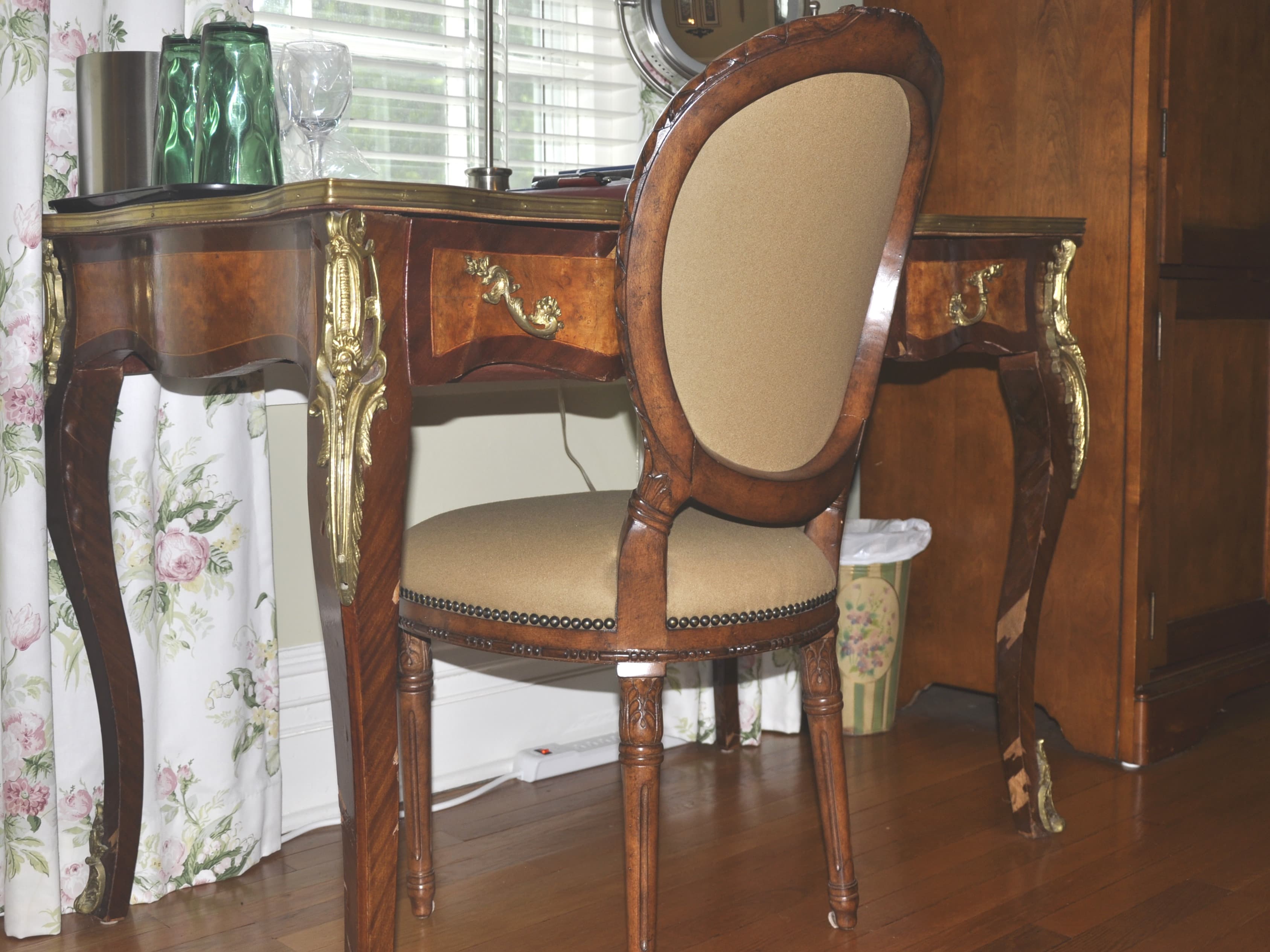 A wooden vanity desk with an ornate chair beside it, set against floral curtains and a wooden wall.
