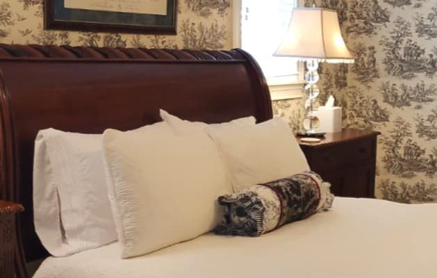 A neatly made bed with white pillows and a decorative throw pillow, against a patterned wallpaper backdrop.